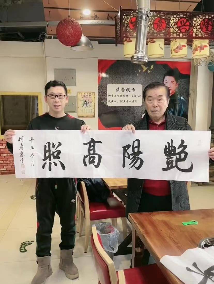 画面中,于小飞和何庆魁一起举着一幅书法作品,上面写着"艳阳高照"四个