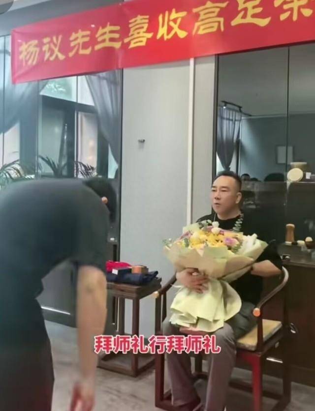 杨毅穿着黑色短t和浅色长裤坐在椅子上,接受弟子的花束,并向老师鞠躬